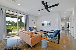 A modern living room with a sofa, a chair, a coffee table, and a TV. There is a ceiling fan, a sliding glass door leading to an outdoor area, and a hallway.