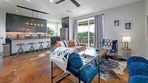Een moderne keuken met barkrukken, een eethoek met een tafel en stoelen, een plafondventilator en een groot raam met gordijnen.
