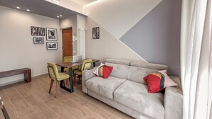 Un salon moderne avec un canapé gris, une table à manger avec des chaises et un ordinateur portable. Des tableaux encadrés ornent le mur et un appareil de climatisation est fixé au plafond.