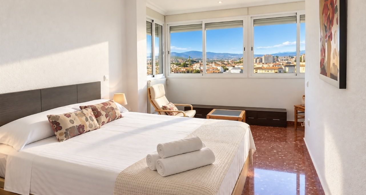 A hotel room with a large bed, a chair, a small table, and a view of the city through large windows.