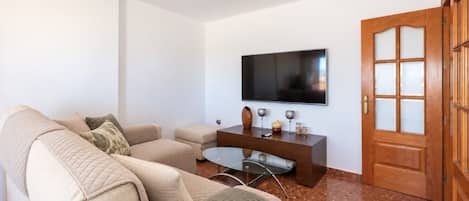 A living room with a beige sofa, a glass coffee table, a flat-screen TV mounted on the wall, and a wooden door with glass panels.