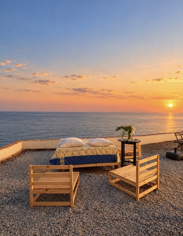 A bed with pillows, a small table with a vase, and two wooden chairs on a pebbled surface by the sea at sunset.