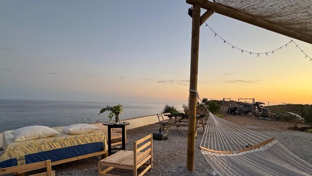 Alojamiento frente a la playa con una hamaca, muebles de madera y vistas al mar al atardecer.