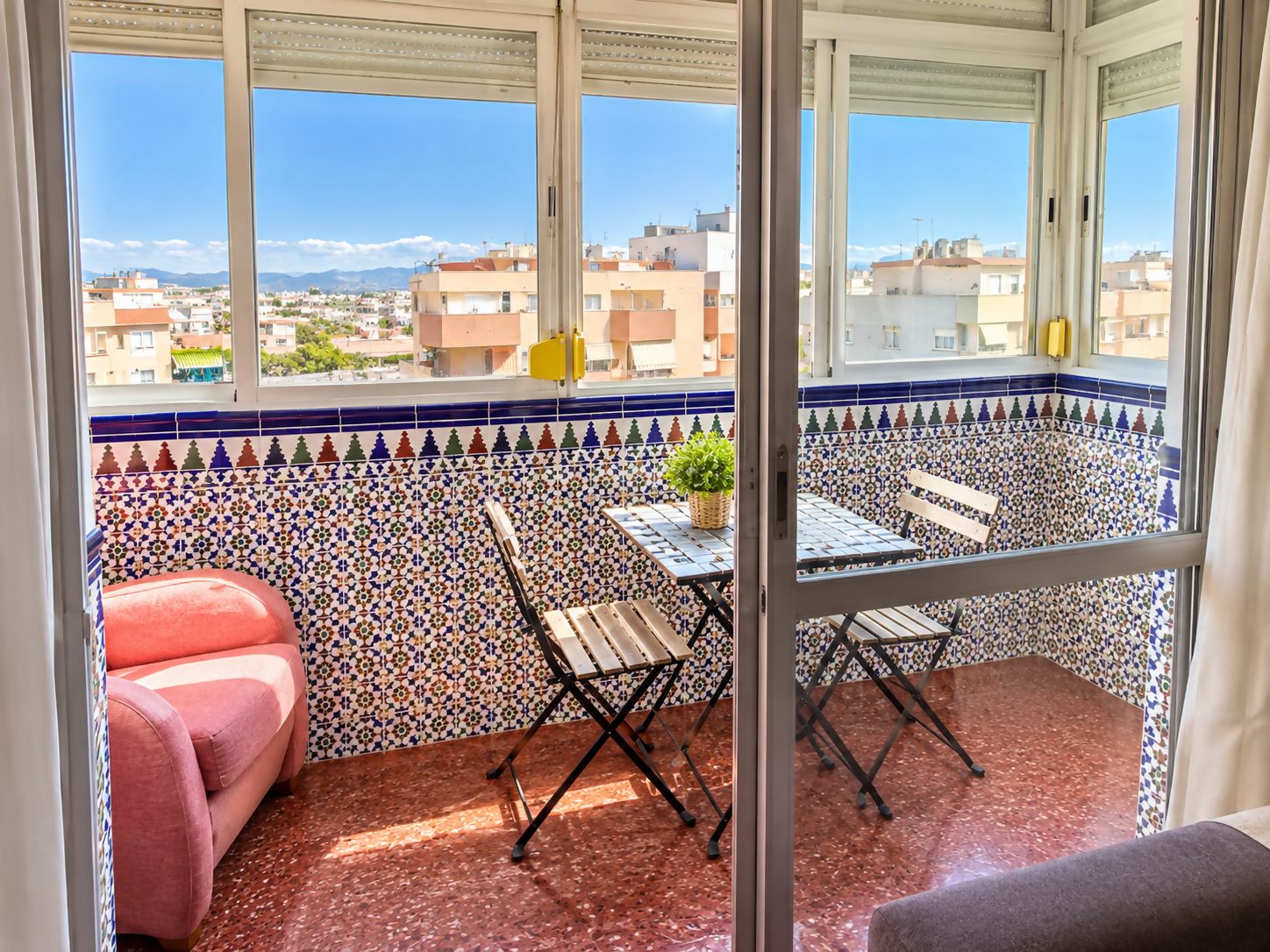 A balcony with a view of the cityscape, a red sofa, a small table with a plant, and a folding chair.