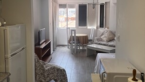 Ein kleines Hotelzimmer mit Küchenzeile, einem Bett, einem Essbereich und einem Fenster mit Jalousien.