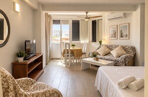 Habitación de hotel con cama, sofá, televisión, mesa de comedor y balcón con cortinas.