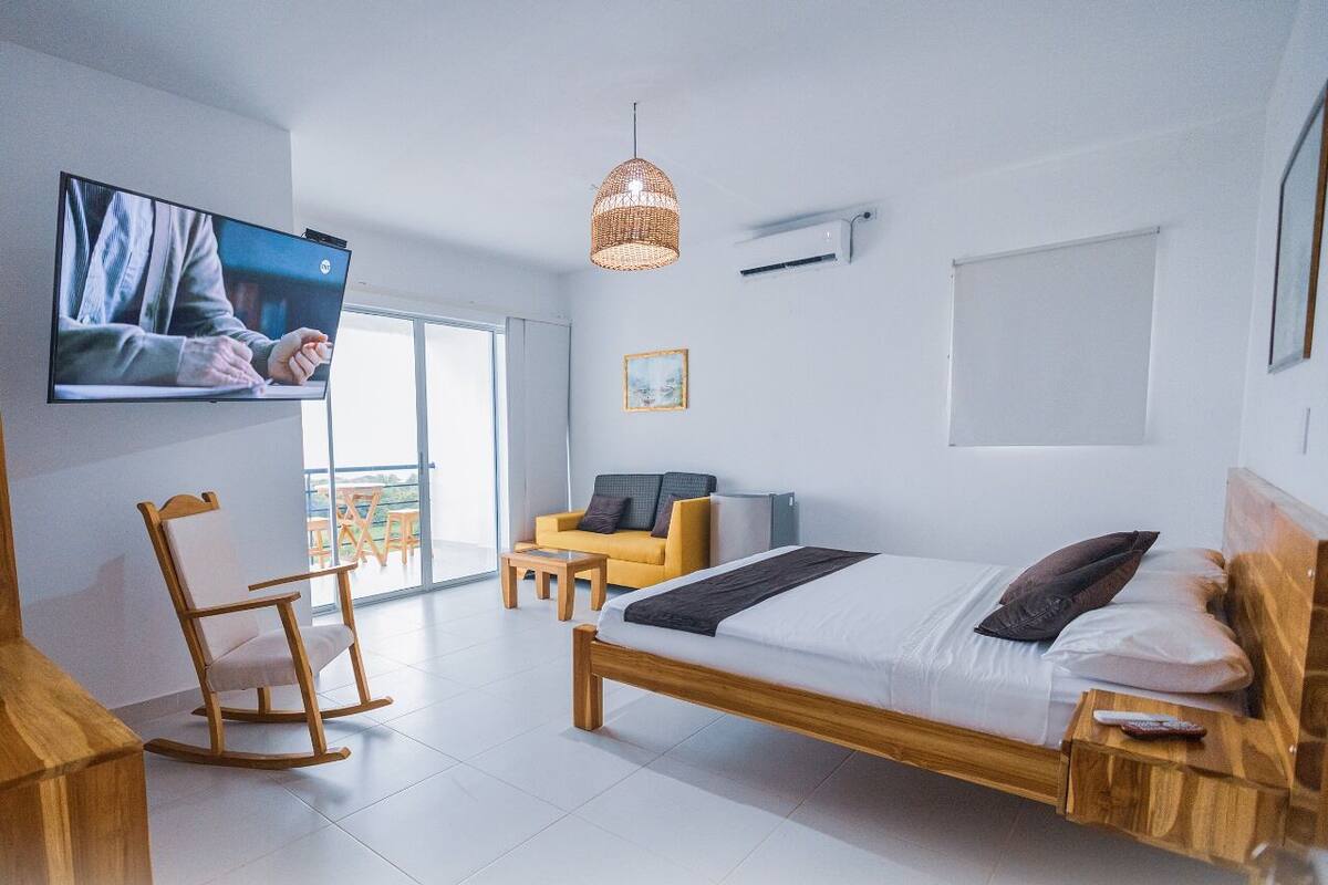 A modern hotel room with a large bed, a TV, a wicker pendant light, a sofa, and a balcony view.