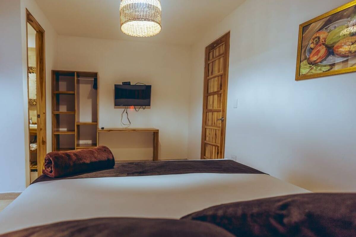 A bedroom with a bed, a TV mounted on the wall, a wooden wardrobe, and a door leading to another room.