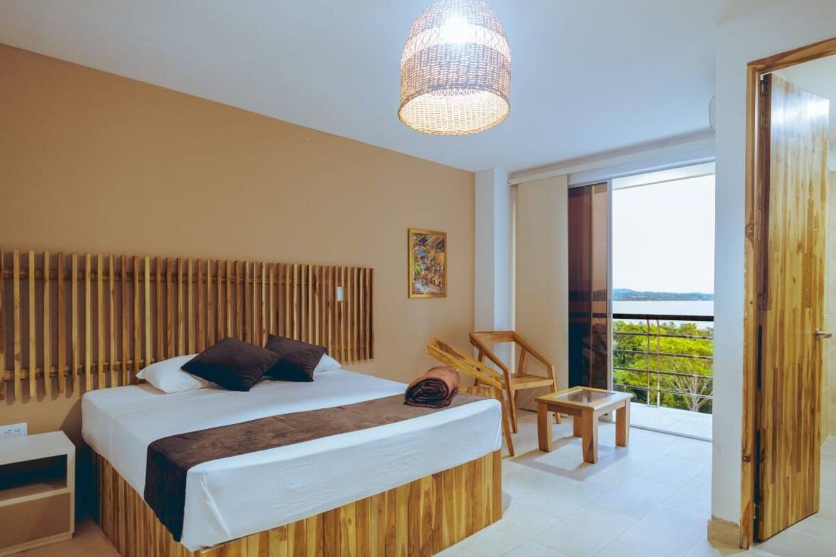 A modern bedroom with a large bed, a wooden headboard, a bedside table, a chair, a small table, and a balcony with a view.