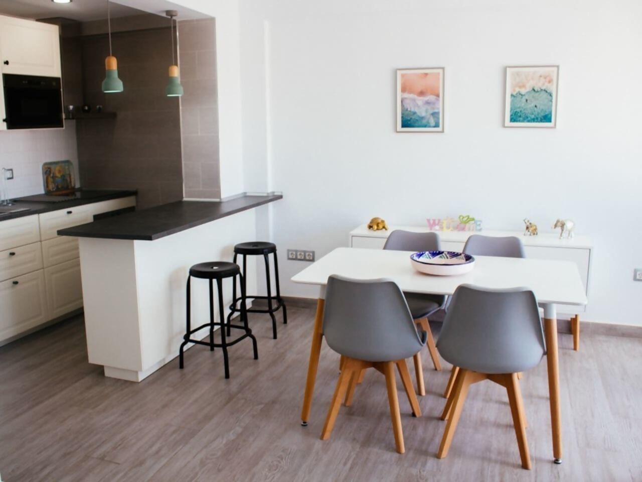 A modern kitchen with a central island, bar stools, a dining table with chairs, and two framed artworks on the wall.