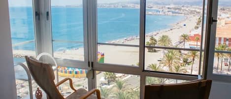 A room with large windows overlooking a beach and ocean. There are two wooden chairs and a white chair.