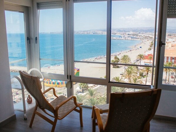 A room with large windows overlooking a beach and ocean. There are two wooden chairs and a white chair.