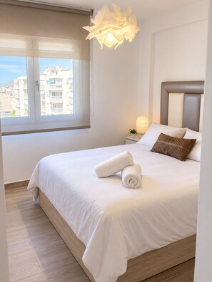 A hotel room with a bed, a window with a view of buildings, a bedside lamp, and a decorative light fixture.