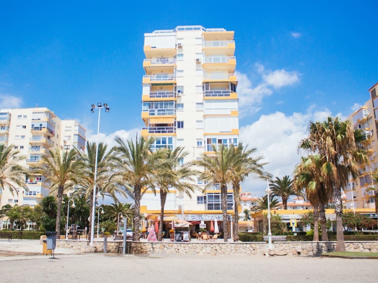Un hôtel à plusieurs étages, doté de balcons, de palmiers et surplombant un ciel d’un bleu limpide.