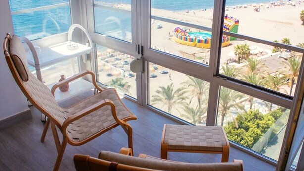 A balcony with wicker chairs and a small table, overlooking a beach with palm trees and a colorful play structure. - Loft in 1st Line Residential with pool (Algarrobo Costa)