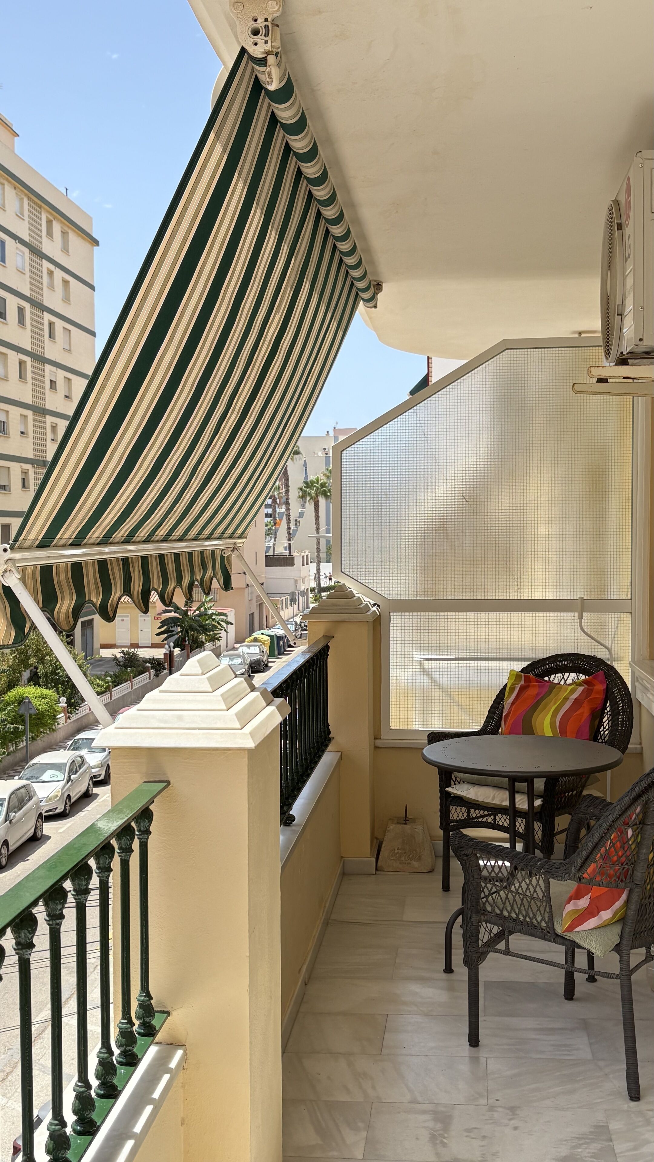 Ein Balkon mit gestreifter Markise, einem Korbstuhl, einem kleinen Tisch und Blick auf die Straße und die Häuser.