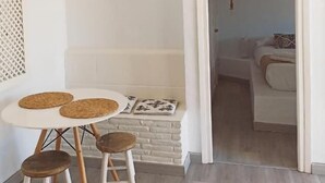 Een compacte woonruimte met een witte muur, een ronde tafel met krukjes, een geometrisch kunstwerk aan de muur, en een doorgang naar een slaapkamer met een bed en een hanglamp.