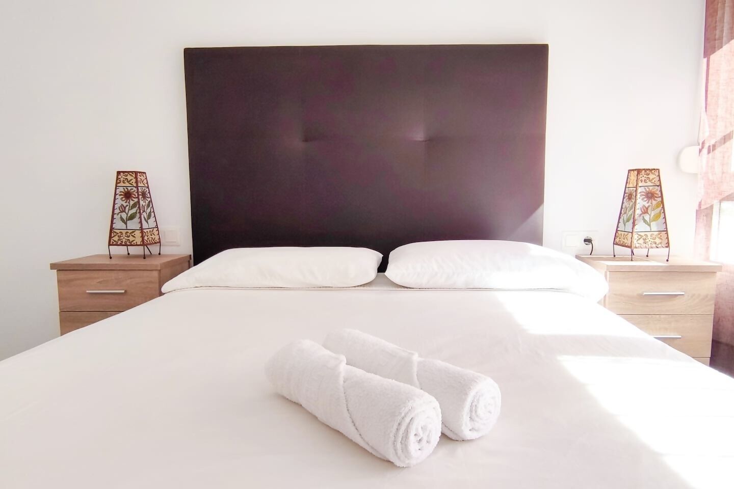 A neatly made bed with white linens and two rolled towels in front. There are two bedside tables with lamps on either side of the bed.