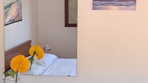 Una habitación de hotel con una cama, un espejo, un cuadro de un atardecer, un jarrón con flores y toallas dobladas sobre un aparador de madera.