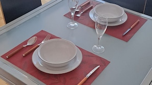 Una mesa de comedor dispuesta con platos, cubiertos y vasos, con un televisor y un sofá al fondo.