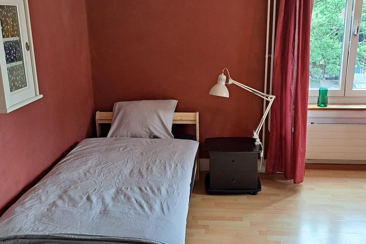A single bed with a white duvet, a bedside table with a lamp, and a window with a red curtain.