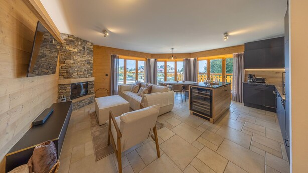 A modern living room with a stone fireplace, a sofa, a coffee table, and a TV mounted on the wall. There is a kitchen area with a countertop and cabinets. - Domaine des Reines C1 - Swimming pool (Haute-Nendaz)