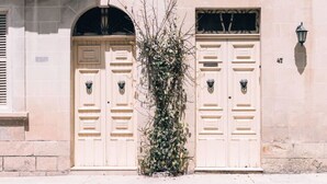 Un bâtiment doté de deux portes symétriques, chacune ornée d’un frappeur en forme de tête de lion. On aperçoit une petite plante devant les portes et un lampadaire sur la droite.