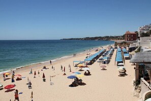 A beach with rows of sunbeds and umbrellas, a clear sky, and buildings in the background.