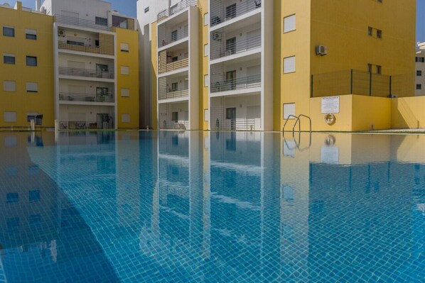 A swimming pool with clear blue water, surrounded by yellow buildings with balconies and windows.