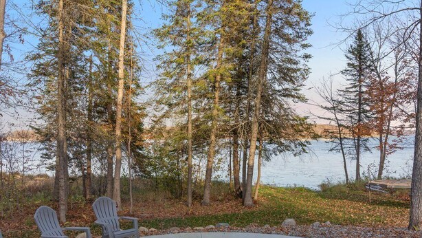 A circular outdoor seating area with a fire pit surrounded by Adirondack chairs, overlooking a body of water and forested area.