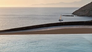 Un paysage côtier avec un voilier sur une eau calme, un mur de pierre et des montagnes au loin.