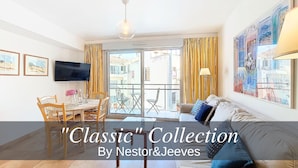 A well-lit living room with a television, a dining area with chairs and a table, a sofa with cushions, and a balcony with curtains. - N&J - VAGUE AZUR - Central - Balcony (Niza)