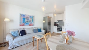 A living room with a sofa, a coffee table, and a dining table with chairs. There is a kitchen in the background with a refrigerator and an oven. - N&J - VAGUE AZUR - Central - Balcony (Niza)