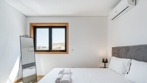 A modern hotel room with a large bed, a bedside lamp, a window with a view, and an air conditioning unit. - GuestReady - Stunning Douro River Vista (Vila Nova de Gaia)