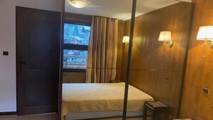 A room with a bed, a small stool, a nightstand, a mirror, and a window with curtains.
