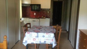A compact living space with a dining table covered by a patterned tablecloth, wooden chairs, a wooden coffee table, and a bed with a plaid blanket. A kitchenette with a microwave and red patterned tiles is visible in the background.