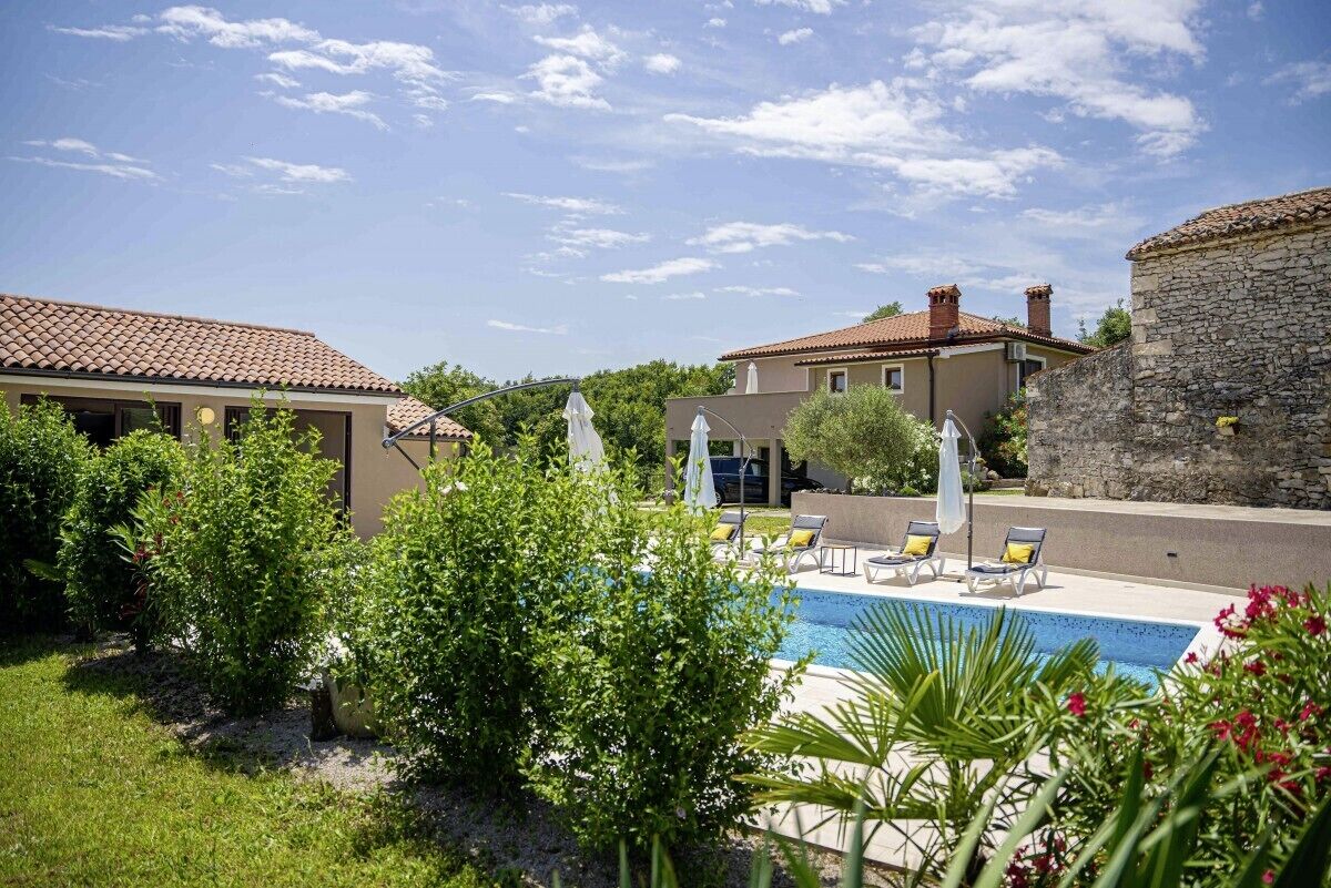 A pool area with lounge chairs and umbrellas, surrounded by greenery and stone buildings.