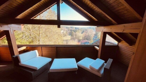 Ein gemütliches Dachzimmer mit schrägen Wänden, Holzbalken und einem großen Fenster mit Blick auf die umliegende Landschaft.