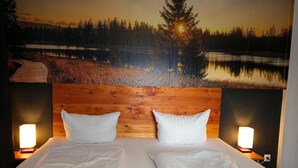 Ein Doppelbett mit weißen Leinentüchern, zwei Kissen und einem hölzernen Kopfteil. Zwei Nachttischlampen und der Blick auf einen Wald und See durch ein Fenster.