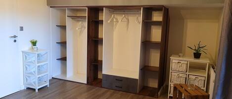 A room with a wooden floor, a white dresser, a wooden wardrobe with multiple shelves, and a small white table with a plant.