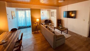 Ein modernes Wohnzimmer mit Holzboden, einer Couch, einem Esstisch und einem an der Wand befestigten Fernseher.