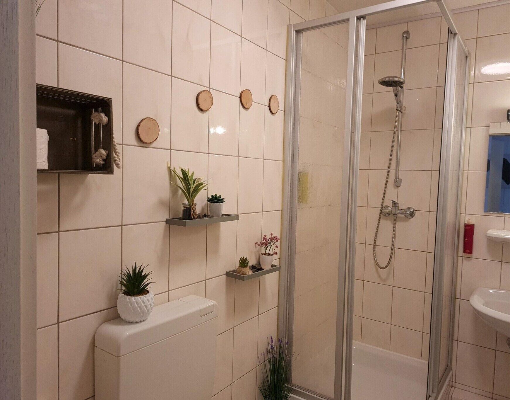 Une salle de bain moderne avec une douche à parois de verre, des toilettes et un lavabo. Des étagères avec des plantes et des objets décoratifs ornent le mur.