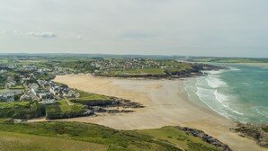 Vista aérea de un pueblo costero con una playa de arena, colinas verdes y un pequeño grupo de casas cerca de la costa.