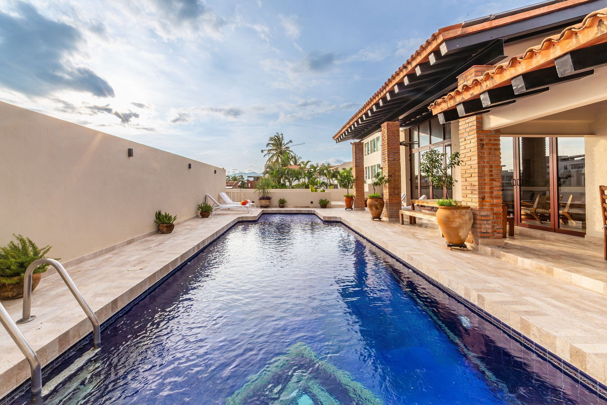 Une piscine avec une terrasse carrelée, entourée d’un bâtiment en briques doté de grandes fenêtres et d’un patio couvert.