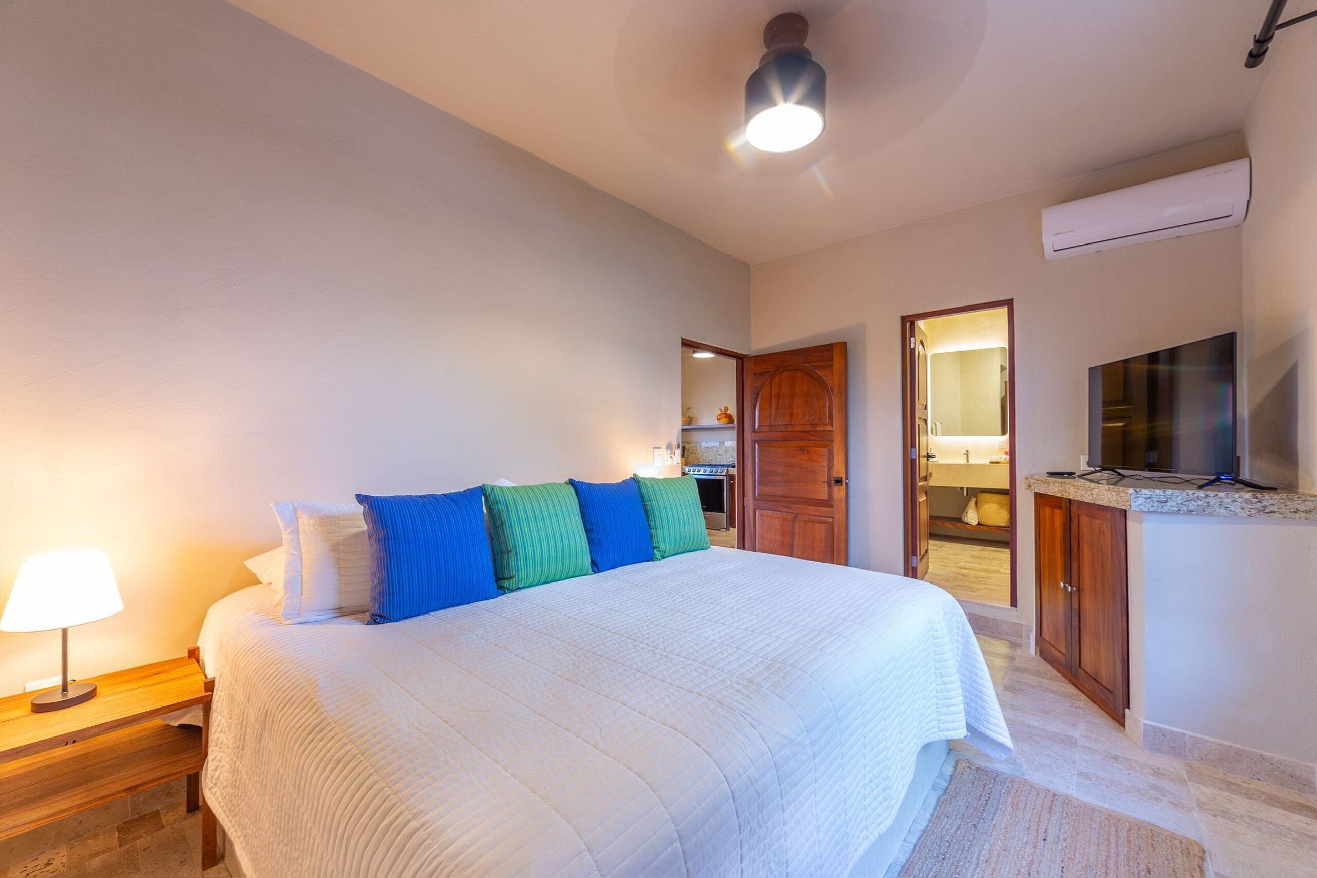 A bedroom with a large bed, wooden nightstands, a desk, a TV, and an air conditioning unit.