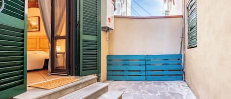 Une ruelle étroite avec un portail bleu, un lit recouvert d’une couverture blanche et un chemin pavé de pierres menant à une porte aux volets verts.