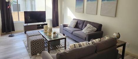 Una sala de estar con un sofá, una mesa de centro y un televisor. Hay cortinas, una lámpara y una ventana.