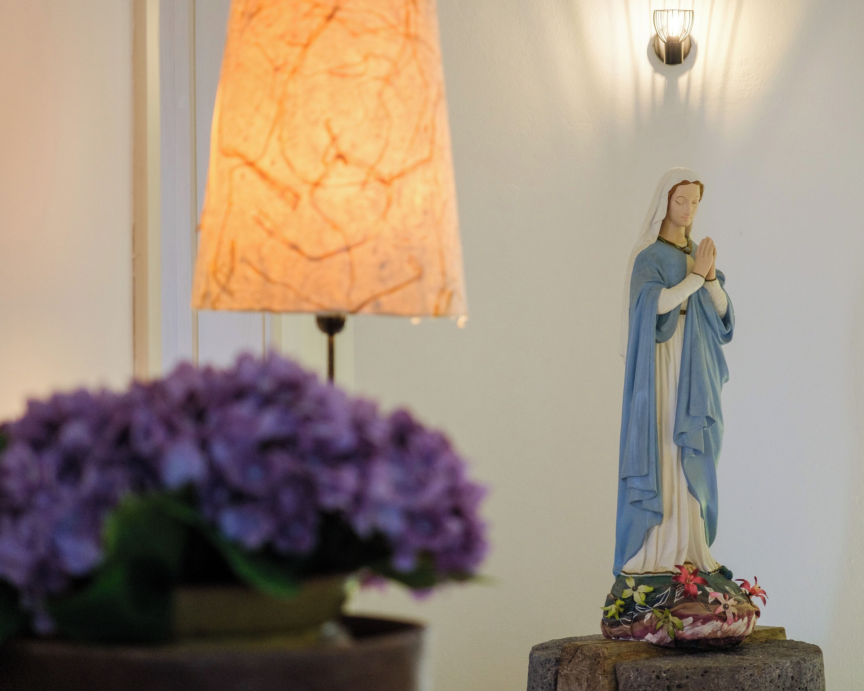 Eine Statue einer religiösen Figur auf einem Sockel, eine Lampe und ein Blumentopf mit violetten Blüten.