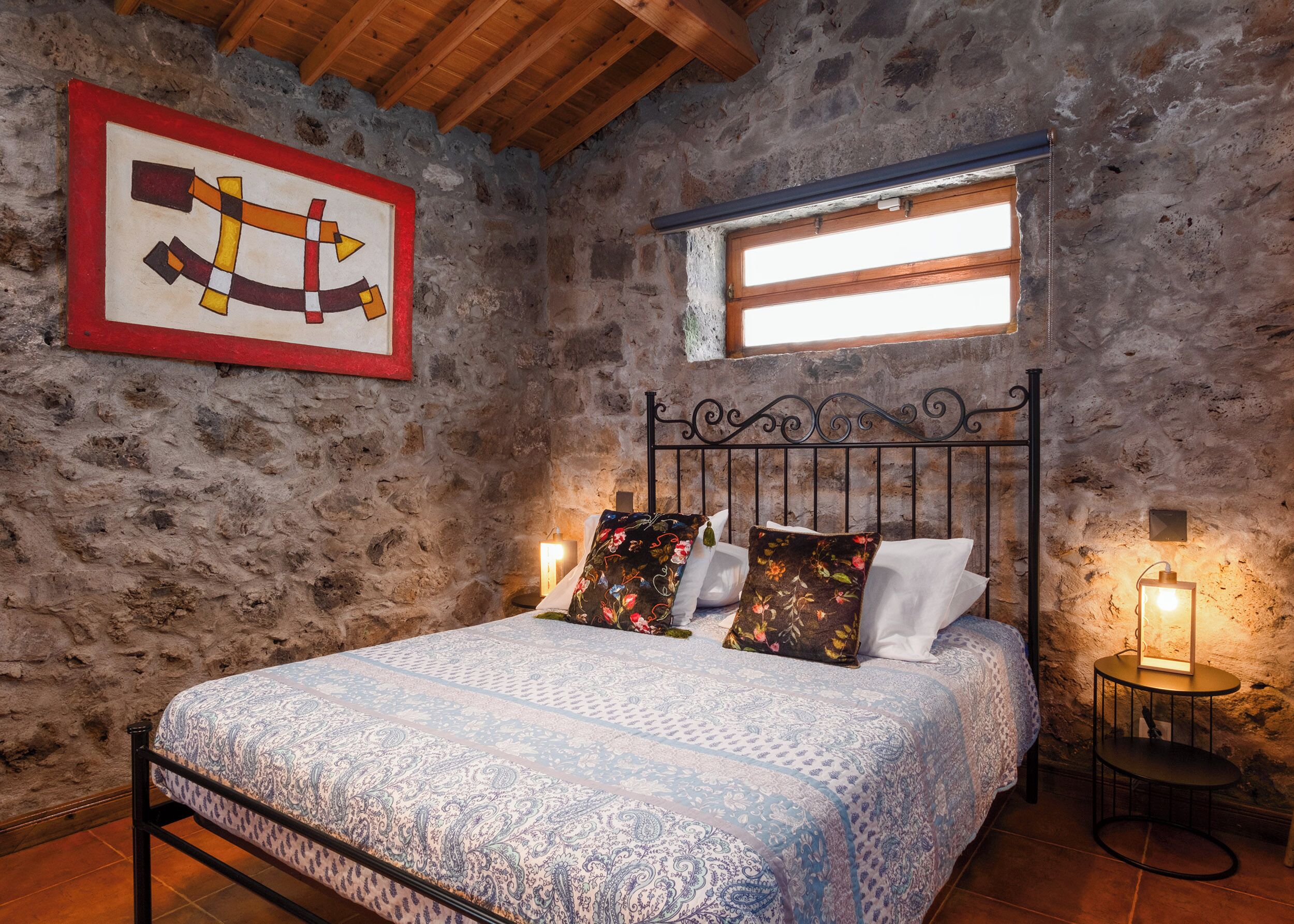 A bedroom with a stone wall, a wooden ceiling, a bed with patterned bedding, a metal headboard, and a window with blinds.