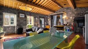 A rustic interior with a stone wall, wooden ceiling beams, a glass dining table, and a leather sofa. - Quinta Morazes - Casa de Campo (Ribeira Grande)